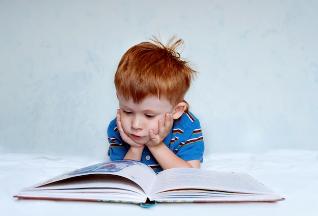 Een schattige kleine jongen met rood haar en een blauw shirt ligt op het bed een boek te lezen