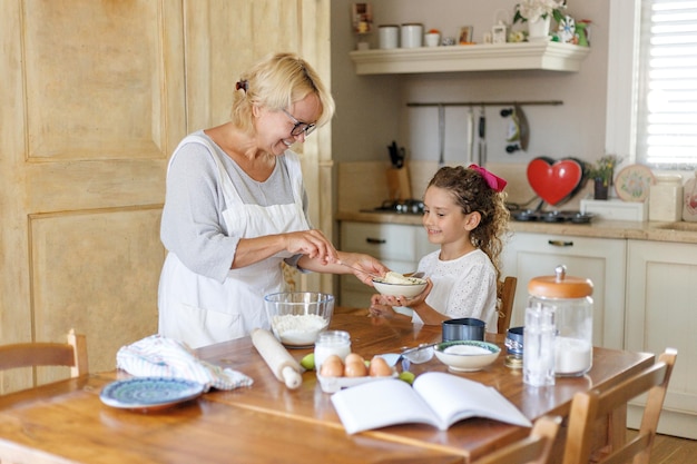 Een schattige oma met kleindochter in de keuken. De kleindochter houdt het deeg in de vorm van een hart, haar grootmoeder kijkt ernaar.