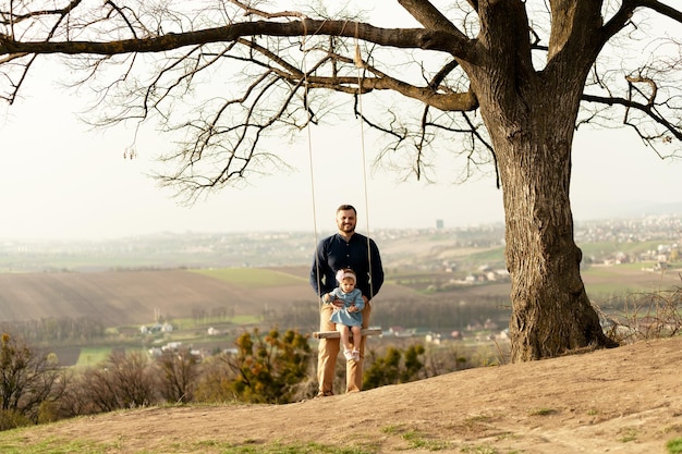 Een schattige vader met een baard speelt met zijn dochtertje op een schommel die op een heuvel staat met uitzicht op de vroege lentespelen met vader