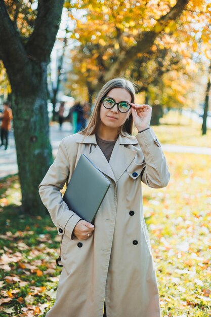 Een schattige vrouw met een bril en lang haar loopt in een herfstpark met een laptop in haar handen werk online in de frisse lucht