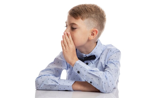 Een schooljongen in een blauw shirt praat fluisterend met zijn hand voor zijn mond. Geïsoleerd op een witte muur. Ruimte voor tekst.