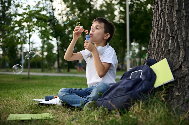 Een schooljongen van de lagere klassen rust in het park, zittend op het groene gras na school, bellen blazend. Schooltas met werkboeken en schoolspullen liggend op het gras