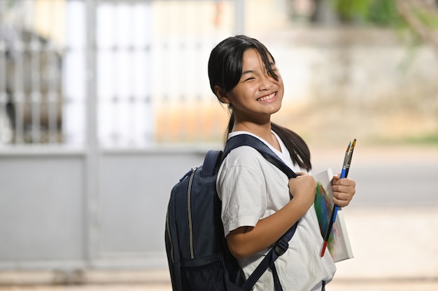 Een schoolmeisje houdt schildersbenodigdheden vast en draagt een schooltas terwijl ze staat te wachten op een schoolbus.