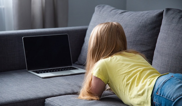 Een schoolmeisje in spijkerbroek en een geel t-shirt op de bank thuis kijken naar een online les op de computer. afstandsonderwijs tijdens het coronavirus