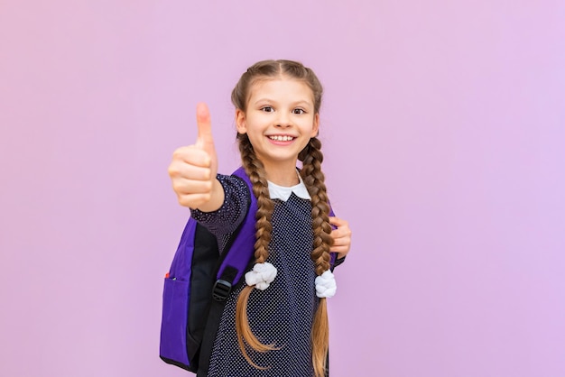 Een schoolmeisje met een koffertje steekt een duim omhoog en glimlacht een klein meisje met een koffertje op een geïsoleerde achtergrond