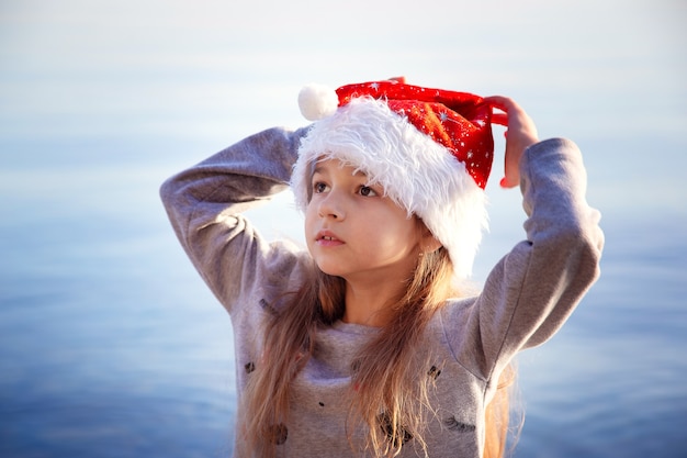 Een schoolmeisje met een nieuwjaarshoed zit aan de kust. Kerst in warme landen. Vakantie en nieuwjaarsvakantie aan zee