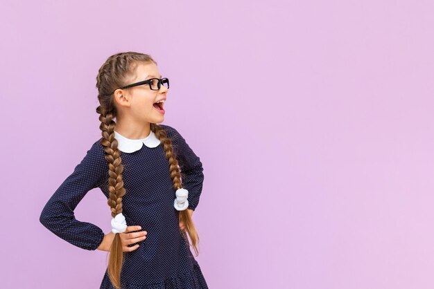 Een schoolmeisje met staartjes houdt haar handen op haar middel en glimlacht op een geïsoleerde paarse achtergrond