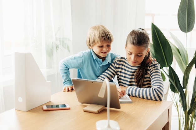Een schoolmeisje zit aan een tafel met haar jongere broer en doet een opdracht op een tablet