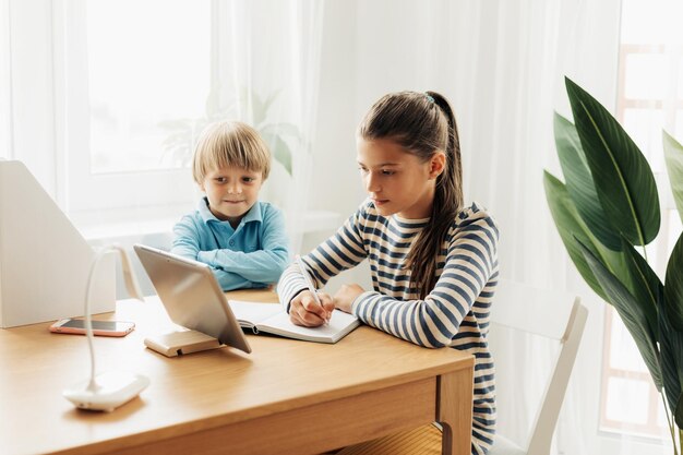 Een schoolmeisje zit aan een tafel met haar jongere broer en doet een opdracht op een tablet