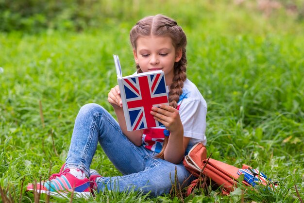 Een schoolmeisje zit op een groen gazon en leest een boek over de Engelse taal