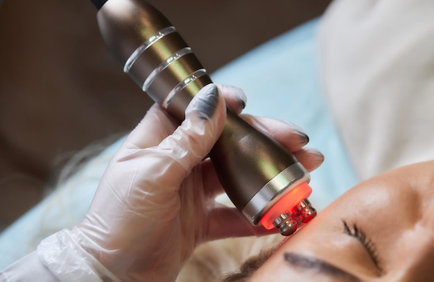 Een schoonheidsspecialist in rubberen handschoenen doet de rflifting-procedure op het gezicht Op de achtergrond het Rf-cosmetologie-apparaat Close-up Zijaanzicht Het concept van professionele cosmetologie en verjonging