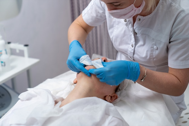 Een schoonheidsspecialiste in een masker en handschoenen maakt een gezichtsreinigingsprocedure voor een meisje met een probleemhuid