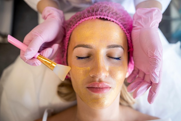 Een schoonheidsspecialiste past een gouden masker met een borstel toe op het gezicht van een jonge mooie vrouw in een schoonheidskliniek Spa salon Schoonheidsspecialist die de procedure van de gezichtshuidverzorging doet