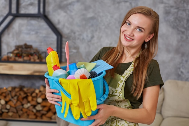 Een schoonmaakster staat in de woonkamer met een blauwe emmer gevuld met chemicaliën en voorzieningen om op te ruimen in haar hand. Schoonmaakdienst.