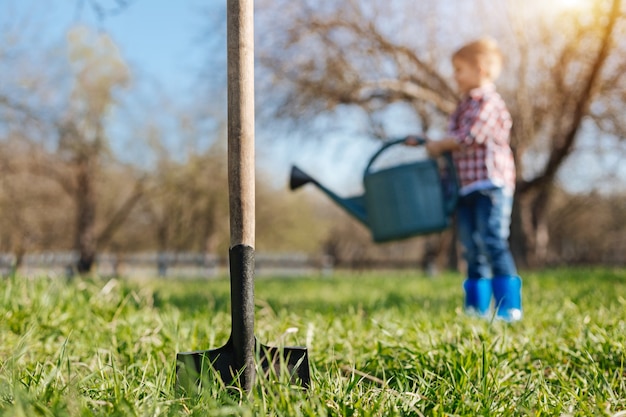 Een schop die grond graaft met een kleine jongen op de achtergrond die een lentegazon met een groene gietende pot water geeft