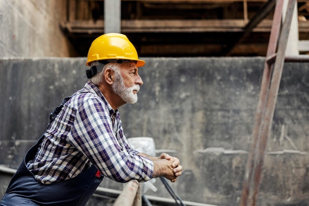 Een senior fabrieksadviseur in overall met een helm op zijn hoofd leunend op de reling