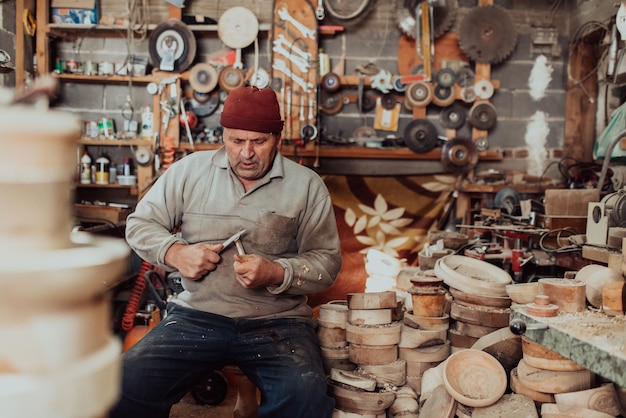 Een senior man zit in de werkplaats en verwerkt houten gebruiksvoorwerpen op de oude handmatige manier.