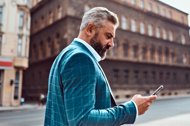 Een senior zakenman in een blauw pak in een stedelijke omgeving met behulp van een smartphone.