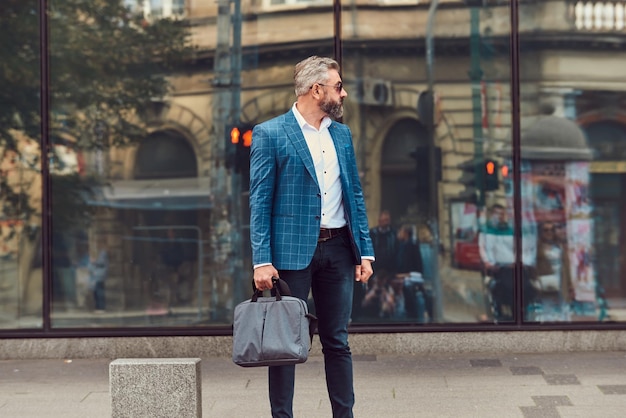 Een senior zakenman in een blauw pak met een koffer die door de stad loopt.