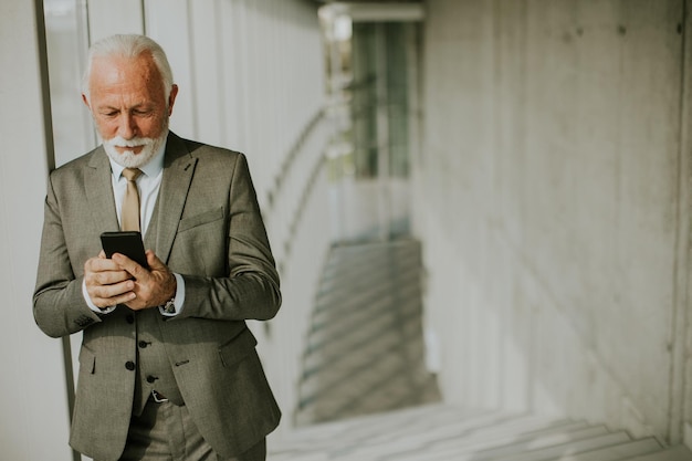 Een senior zakenman staat in een kantoorgang gefocust op zijn mobiele telefoon