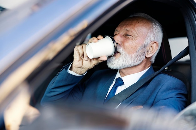 Een senior zakenman zit in de ochtendspits in zijn auto en drinkt koffie op weg naar zijn werk