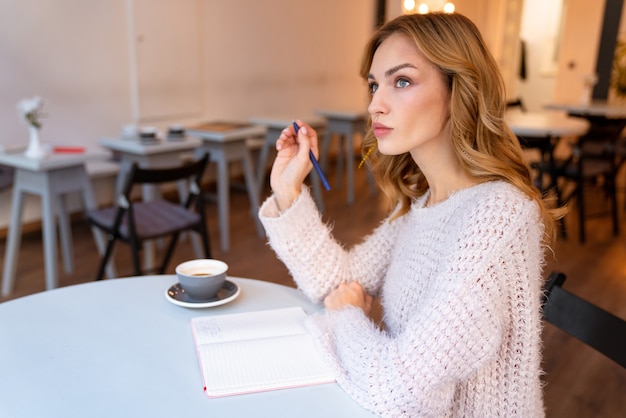 een serieus denkende mooie jonge blonde vrouw zit in café binnenshuis en schrijft notities in een notitieblok.