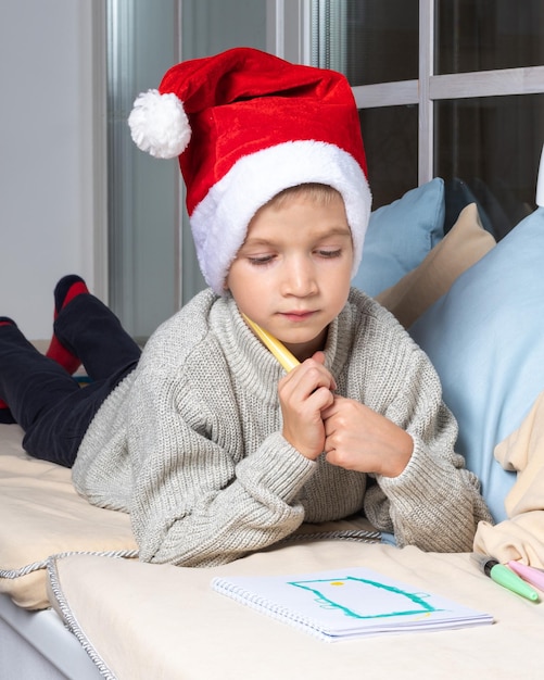 Een serieus gefocust kind met een kerstmuts schrijft thuis een verlanglijstje of een brief aan de kerstman. De jongen tekent een telefoon