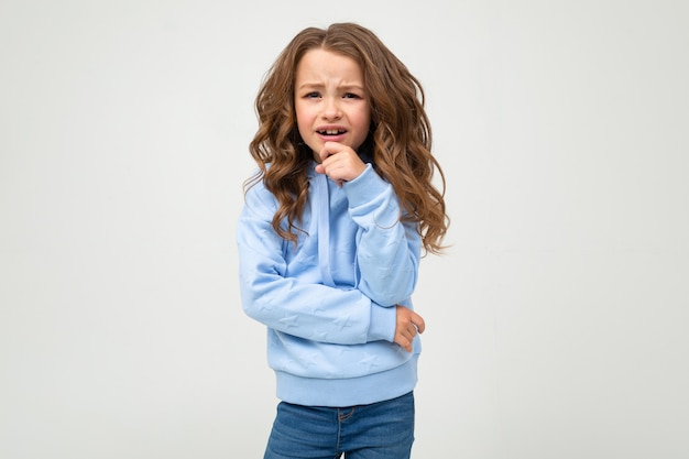 Een serieus meisje in een casual blauwe hoodie staat met een eerlijke uitdrukking terwijl ze een vinger voor haar mond houdt op een witte muur met lege ruimte