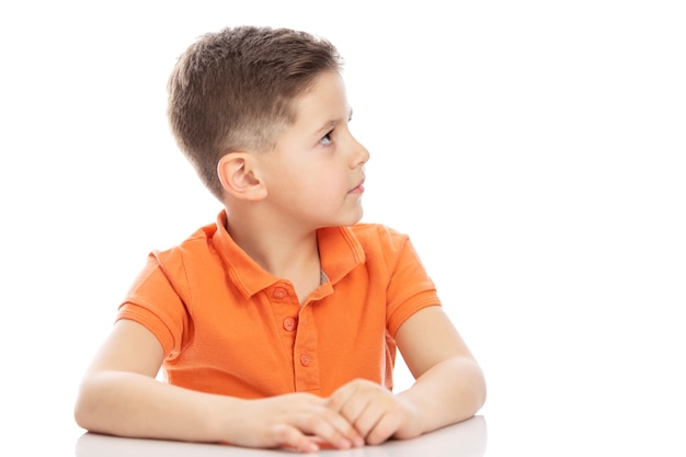 Een serieuze schoolgaande jongen in een fel oranje polo t-shirt zit aan een tafel en kijkt opzij. Isolirvoan op een witte achtergrond.