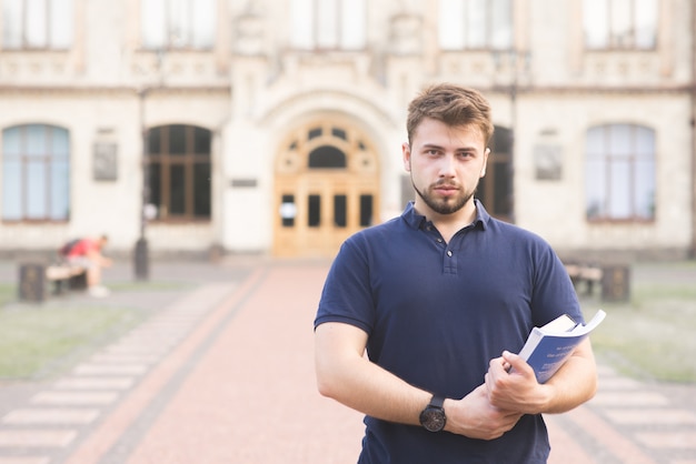 Een serieuze student staat met boeken in handen van het universiteitsgebouw en kijkt naar de camera.