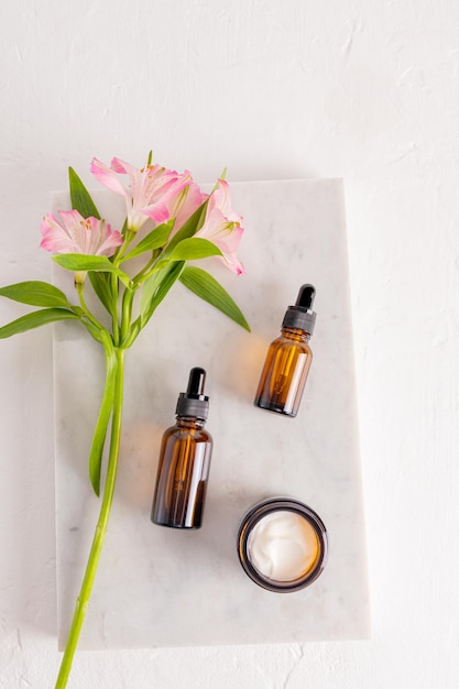 Een serum in twee cosmetische flessen met een druppelaar en een open pot met vochtinbrengende crème voor de huidverzorging van gezicht en lichaam liggen op een marmeren podium Top view Bloemen