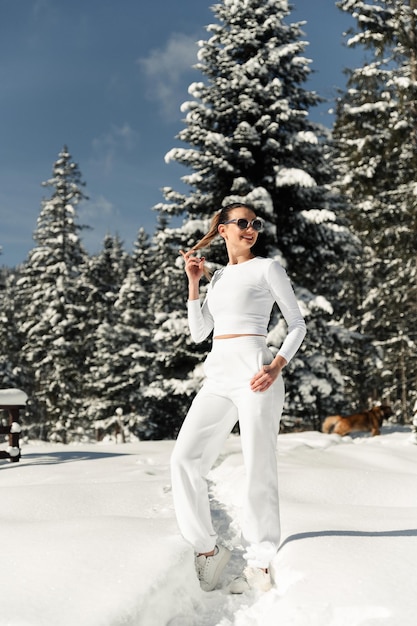 Een sexy meisje in witte kleren en zonnebril staat in een sneeuwjacht tegen de achtergrond van een bos wintervakantie portret van een schattig meisje in de winter