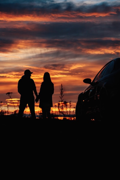 Een silhouet van een paar dat in de buurt van een auto staat tegen de achtergrond van een prachtige zonsondergang of zonsopgang.