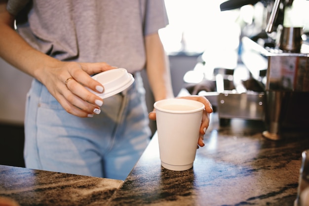 Een slank jong mooi meisje, gekleed in casual outfit, legt voorzichtig het deksel op de koffie in een gezellige coffeeshop. .