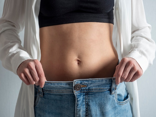 Een slanke vrouw met een blote buik laat zien hoe ze is afgevallen. Jeans zijn te groot geworden. Het concept van voeding en gezond eten