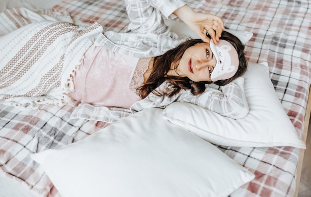 Een slaperig brunette meisje ligt in bed met een blinddoek en wordt 's ochtends wakker. Gezellige huisochtend in schattige pyjama's. Veel plezier, ontspan en droom. Gezonde slaap.