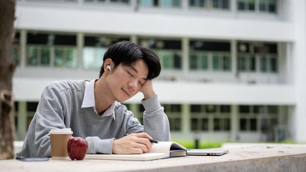 Een slaperige en ontspannen Aziatische mannelijke universiteitsstudent geniet met gesloten ogen van de muziek in een park