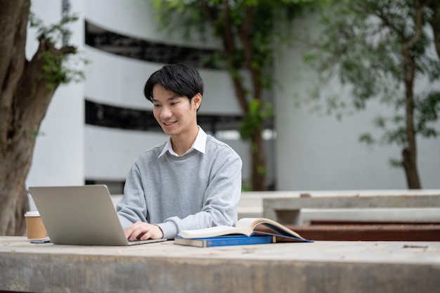 Een slimme Aziatische mannelijke student werkt op afstand in een park aan zijn project op zijn laptop