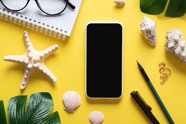 Een smartphone bril een blanco notitieboekje en Monstera Deliciosa bladeren en schelpen op een gele achtergrond Bloggen in de zomer op de werkplek is een bedrijf