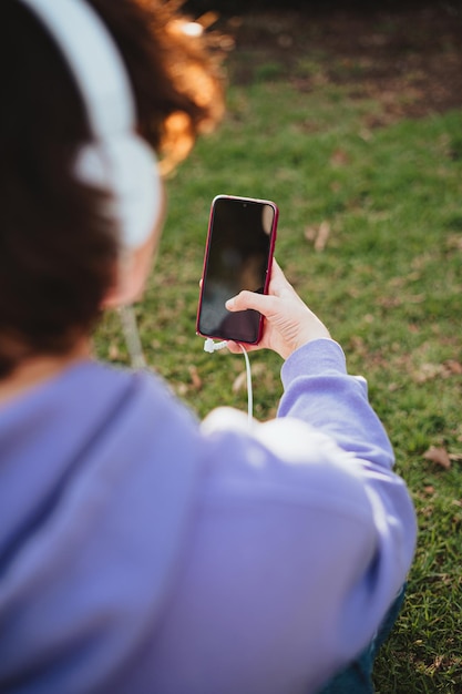 Een smartphone die wordt gebruikt door een tienermeisje. Ze draagt een witte koptelefoon terwijl ze in het gras zit.