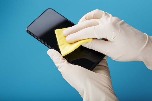 Een smartphone schoonmaken met een steriel geel servet in rubberen handschoenen op een blauwe achtergrond.