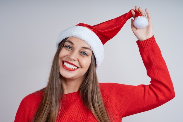 Een speels jong meisje in een rode trui en kerstmuts. Studio die op grijze achtergrond is ontsproten.