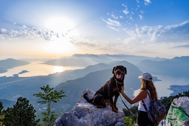 Een sportief meisje met een rugzak staat op de rand van een berg met een rottweilerhond