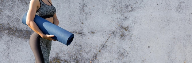 Een sportief slank meisje in een legging en een top staat bij een betonnen muur met een trainingsmat die tussen de oefeningen rust