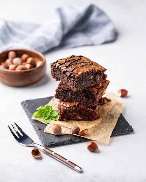 Een stapel chocolade brownies met munt hazelnoten en gezouten caramel op bakpapier op een lichte achtergrond met vork en schaal met noten