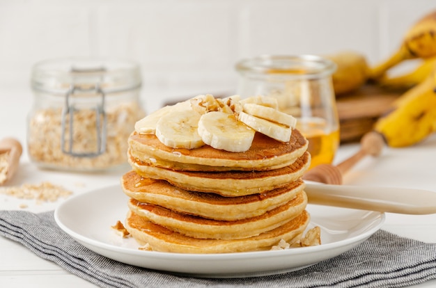 Een stapel havermout banaan pannenkoeken met plakjes verse bananen, walnoten en honing bovenop met kopje thee op een witte houten achtergrond. Een gezond ontbijt. Kopieer ruimte.