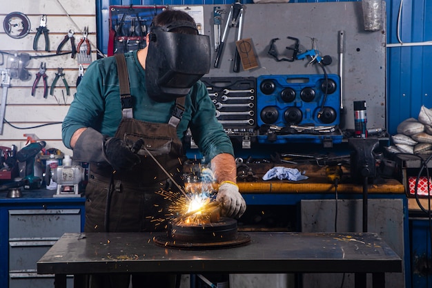 Een sterke man is een lasser in een zwart T-shirt, in een lasmasker en lassersleer, een metalen product wordt gelast met een lasmachine in de garage, gele vonken vliegen naar de zijkanten