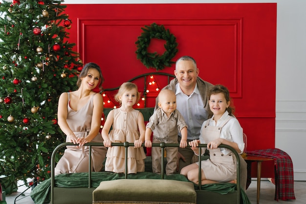 Een stijlvol gezin met drie kinderen zit op een bed bij een kerstboom. Een gezellige kerstvakantie