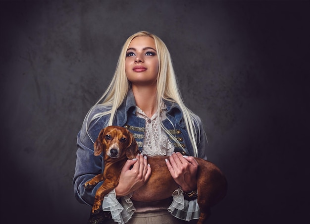 Een stijlvolle blonde vrouw gekleed in een ouderwets jasje houdt een rode dassenhond vast.