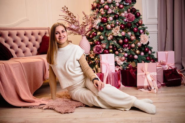Een stijlvolle, gelukkige jonge vrouw in een modieus wit gebreid pak zit op een zacht vloerkleed in een gezellige kamer bij een kerstboom Modieus meisjes vrolijk model rust in de kerststudio